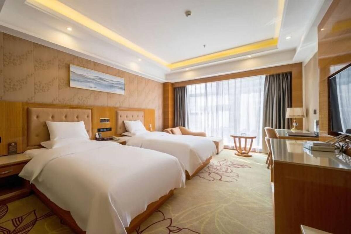 A hotel room with two beds, a desk, and a large window with curtains.