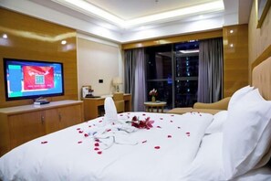 Quarto de hotel com uma cama grande, televisão, escrivaninha e vista da cidade à noite.