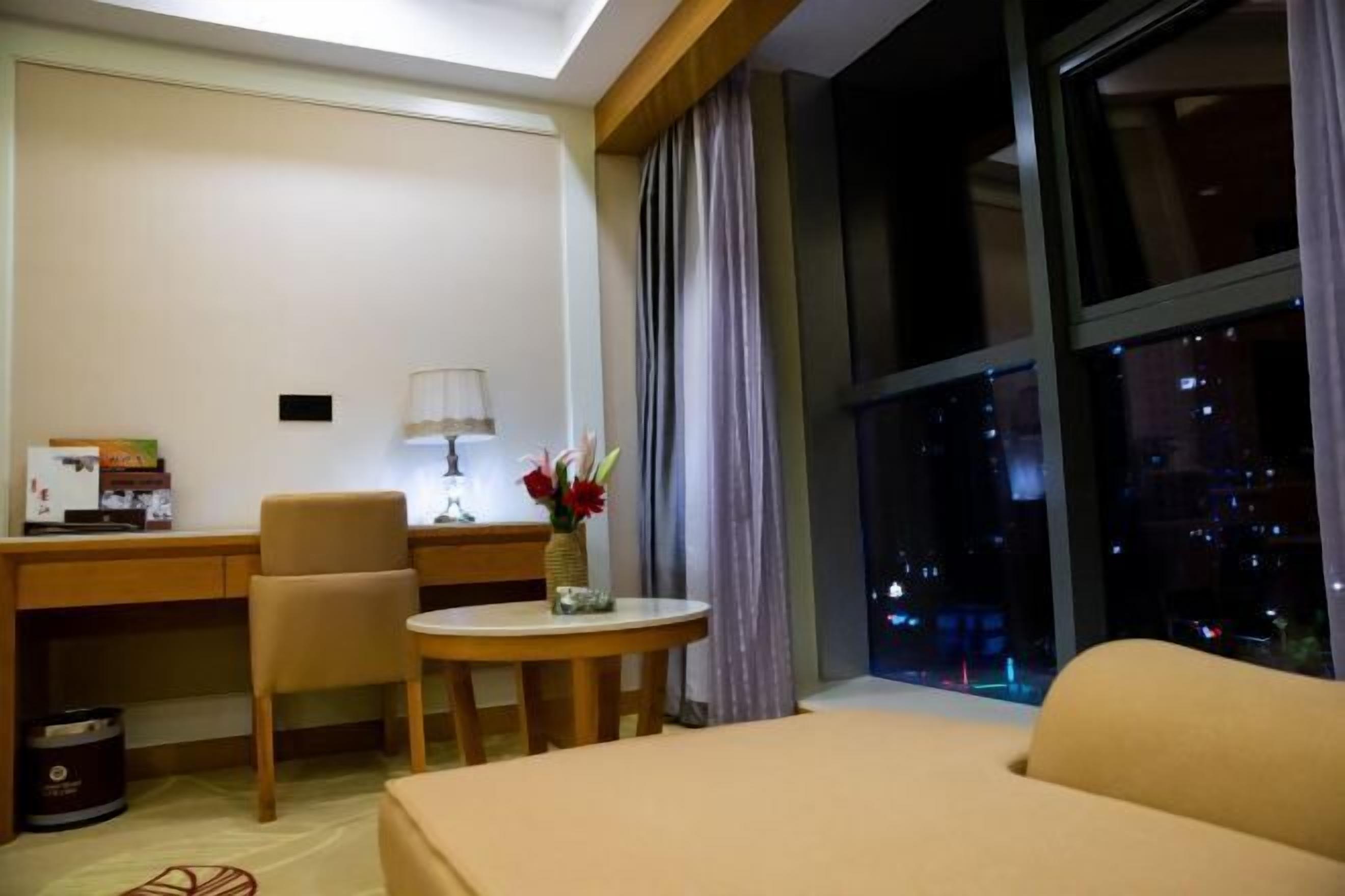 Ett hotellrum med en säng, ett skrivbord, en stol, ett litet bord med en vas med blommor och ett fönster med utsikt över staden på natten.