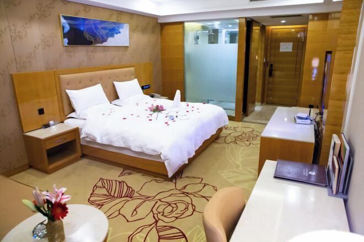 Quarto de hotel com cama, mesas de cabeceira, escrivaninha, cadeira, um vaso com flores e um quadro na parede.