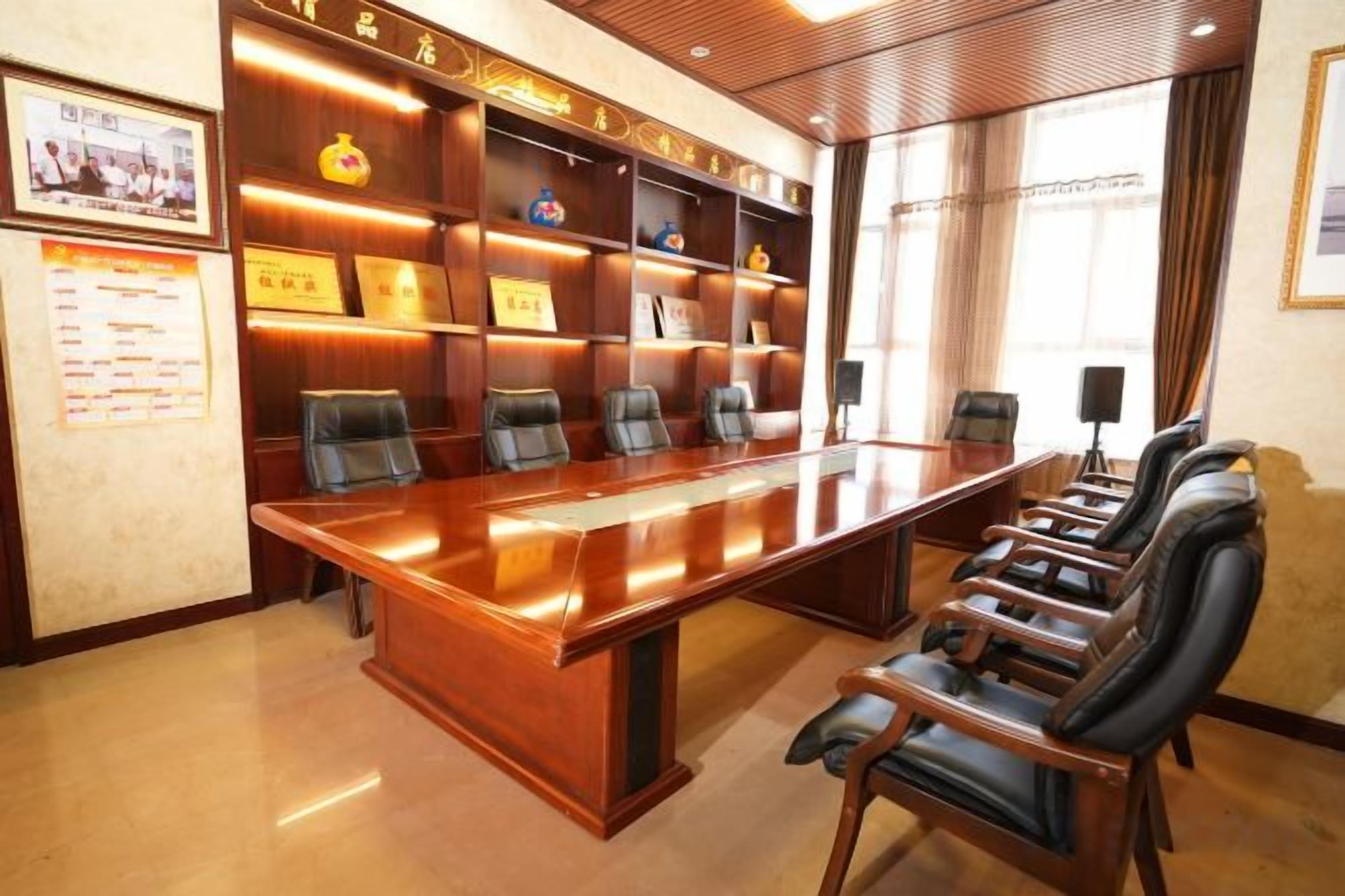 A conference room with a long table and multiple chairs, a bookshelf with awards, and a window with curtains.