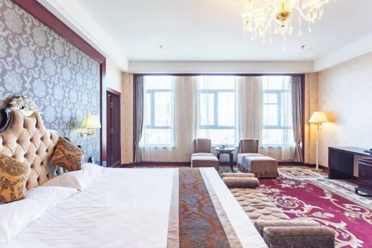 A hotel room with a large bed, a chandelier, a desk, and a view of the city through large windows.