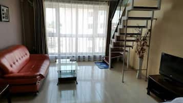 A living room with a red sofa, a glass coffee table, a TV on a stand, a staircase, and a large window with curtains.