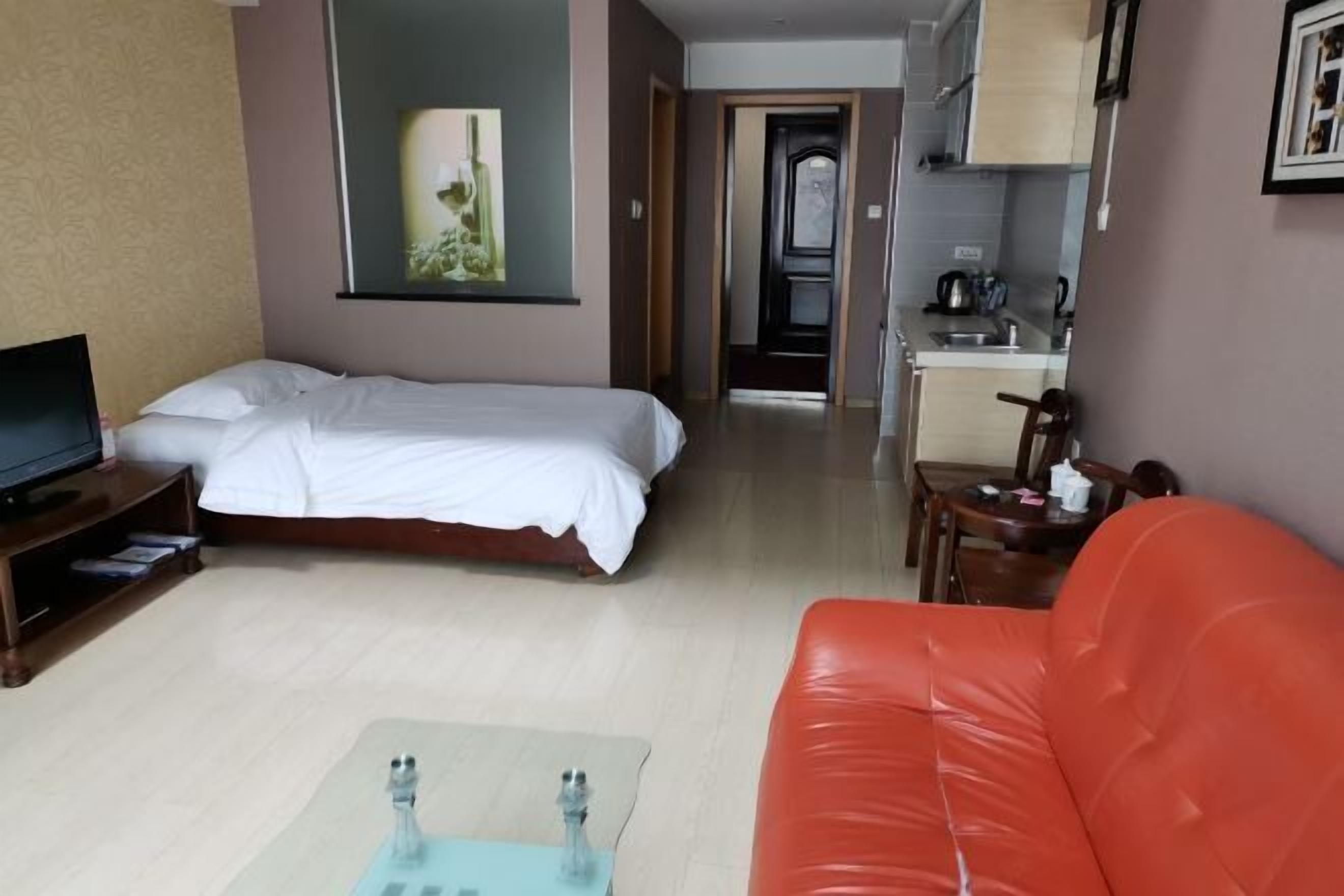 Una habitación de hotel con una cama, televisión, sofá rojo, una mesita y una zona de cocina al fondo.