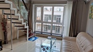 A modern living room with a glass coffee table, a beige sofa, a staircase with glass railings, and a large window with curtains. - Fubang Hotel Apartment (Suifenhe)