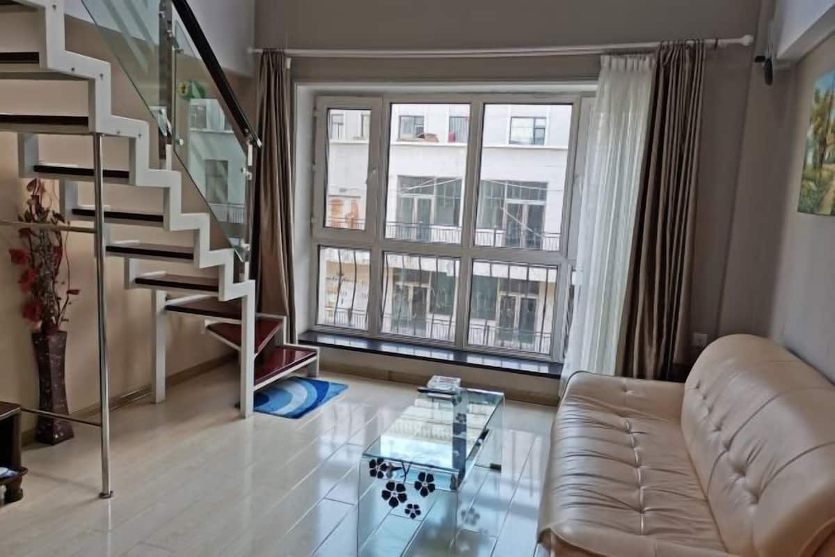 A modern living room with a glass coffee table, a beige sofa, a staircase with glass railings, and a large window with curtains.
