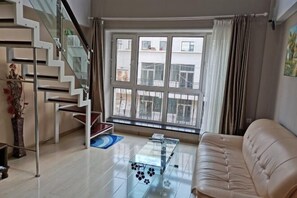Una sala de estar moderna con una mesa de centro de vidrio, un sofá color beige, una escalera con barandillas de cristal y un ventanal con cortinas.