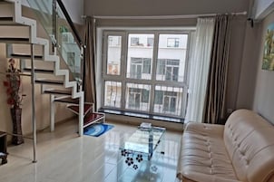 A modern living room with a glass coffee table, a beige sofa, a staircase with glass railings, and a large window with curtains.