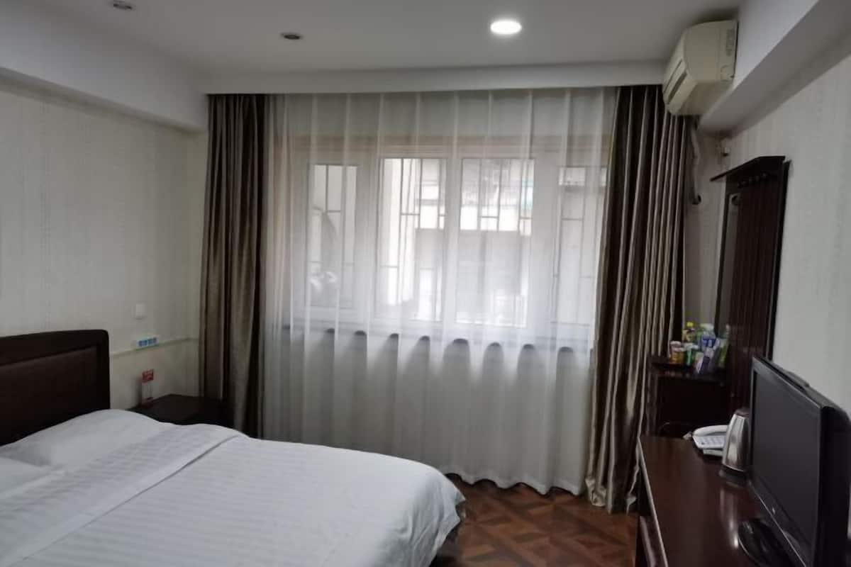 A hotel room with a bed, a desk with a computer, a chair, a window with curtains, and an air conditioning unit.