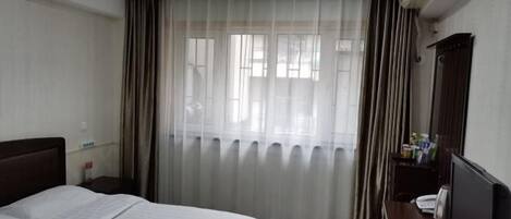 A hotel room with a bed, a desk with a computer, a chair, a window with curtains, and an air conditioning unit.