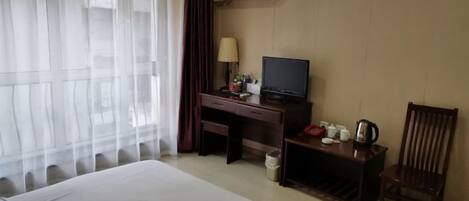 A hotel room with a bed, a desk with a television, a chair, and a small table with cups and a kettle.