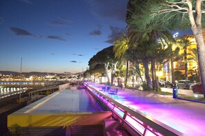 Una passeggiata lungomare con panchine e palme, che si affaccia su uno specchio d'acqua con le luci della città sullo sfondo.