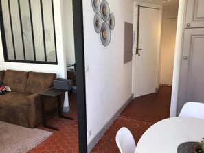 Un salon avec un canapé marron, une petite table et une chaise blanche. On aperçoit un élément décoratif mural et une porte à l’arrière-plan.