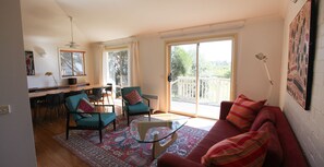 Ein großzügiger Wohnbereich mit einem roten Sofa, blaugrünen Sessel und einem Glastisch. Es gibt einen Essbereich mit einem Holztisch und Stühlen, sowie einen Balkon mit Blick ins Grüne.