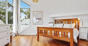 Un dormitorio con cama de madera, cómoda blanca y un ventanal con vistas a árboles.