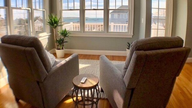 A room with two armchairs, a small round table, and a large window offering a view of the beach and ocean. - Offers  ocean Views and Access & is Great for All Ages! (2249) (West Yarmouth)