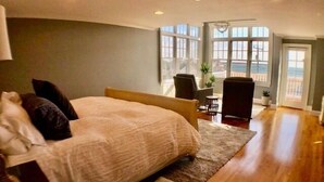 A bedroom with a bed, two chairs, a small table, and a view of the outdoors through large windows. - Offers  ocean Views and Access & is Great for All Ages! (2249) (West Yarmouth)