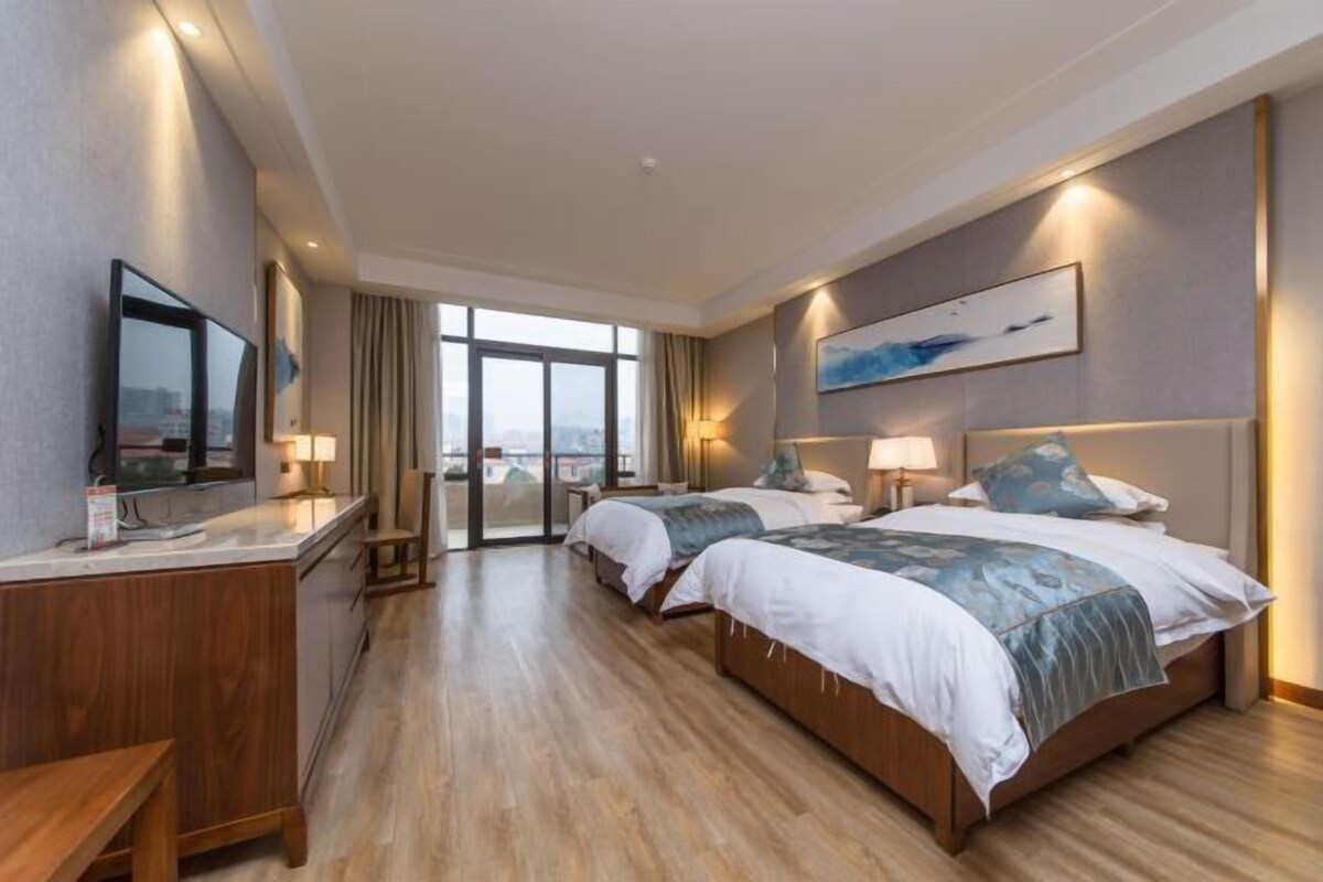 A hotel room with two beds, a desk, and a large window offering a city view.