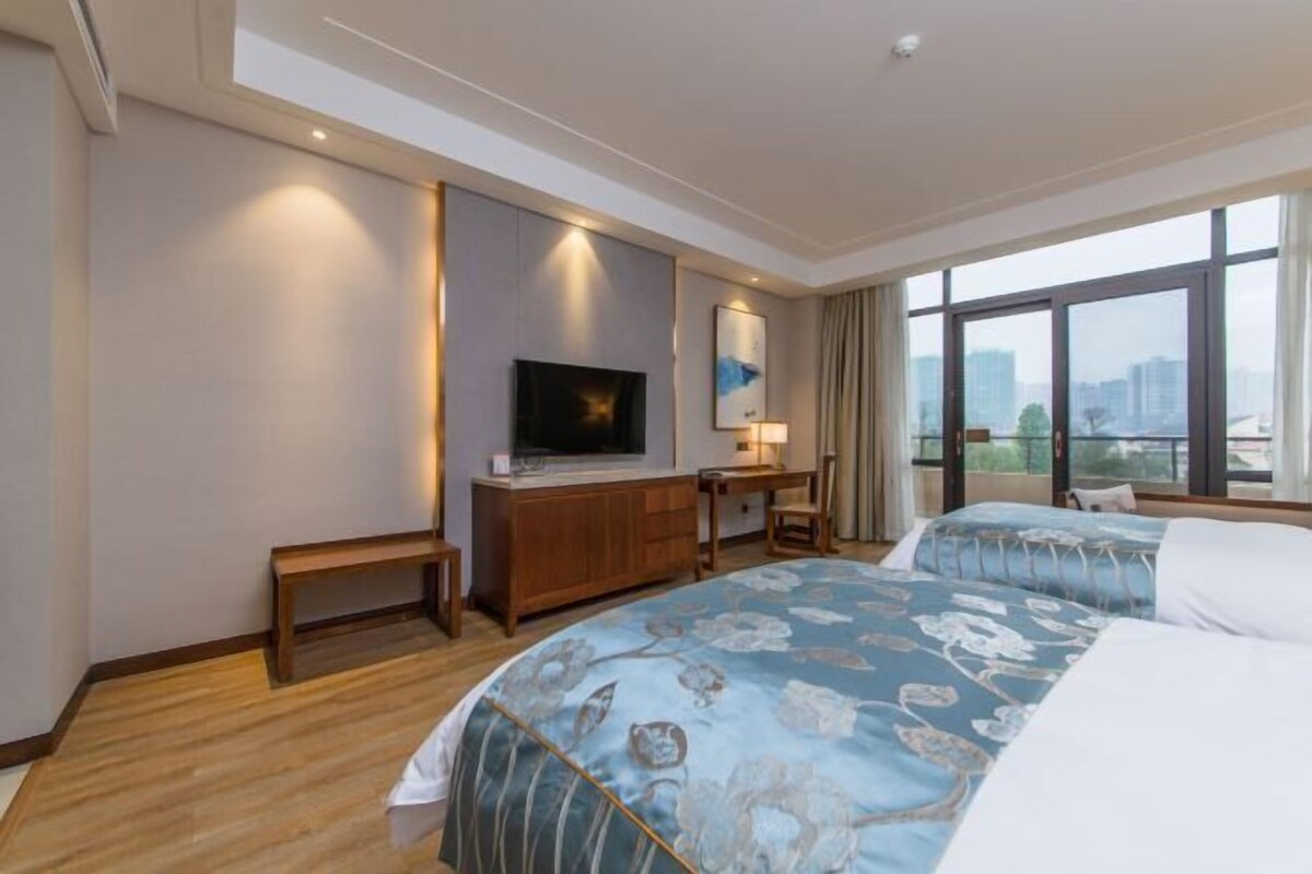 Ein Hotelzimmer mit einem großen Bett, einem Schreibtisch mit Stuhl, einem Fernseher und Blick auf die Stadt durch große Fenster.