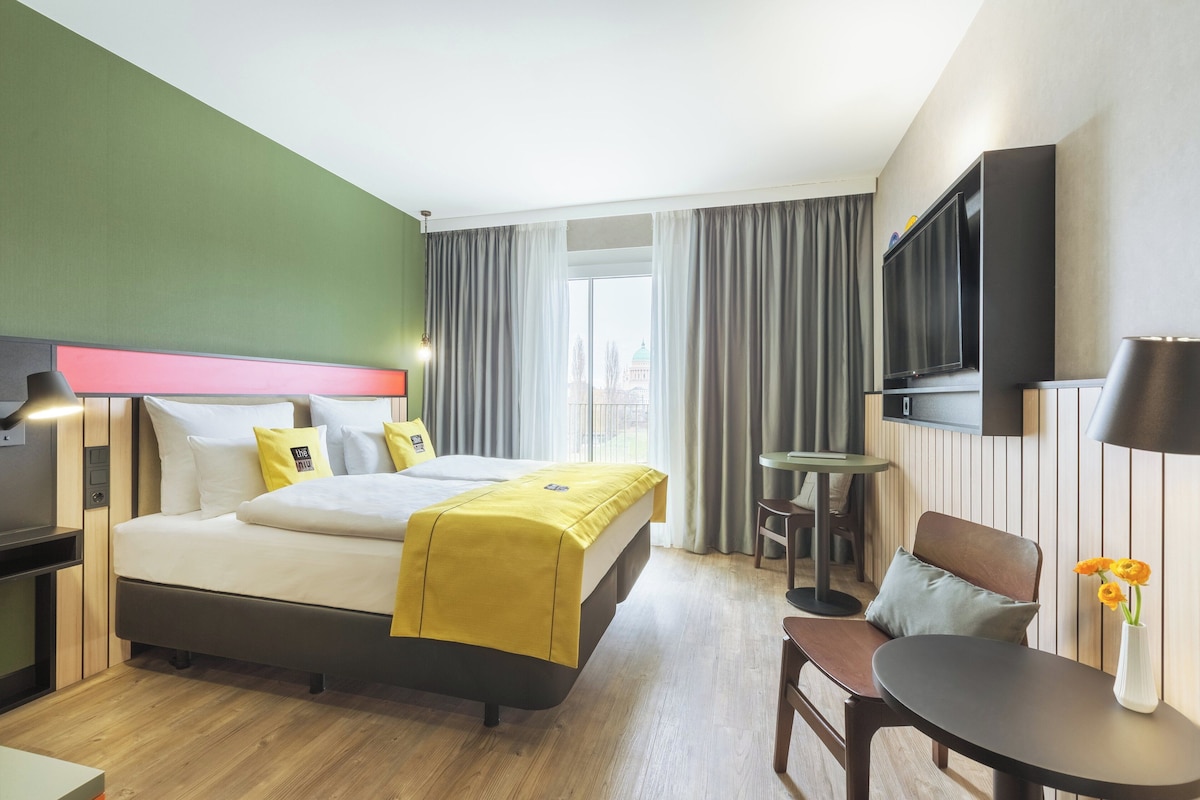Une chambre d’hôtel avec un lit, un bureau, une chaise, une télévision et une fenêtre avec des rideaux.