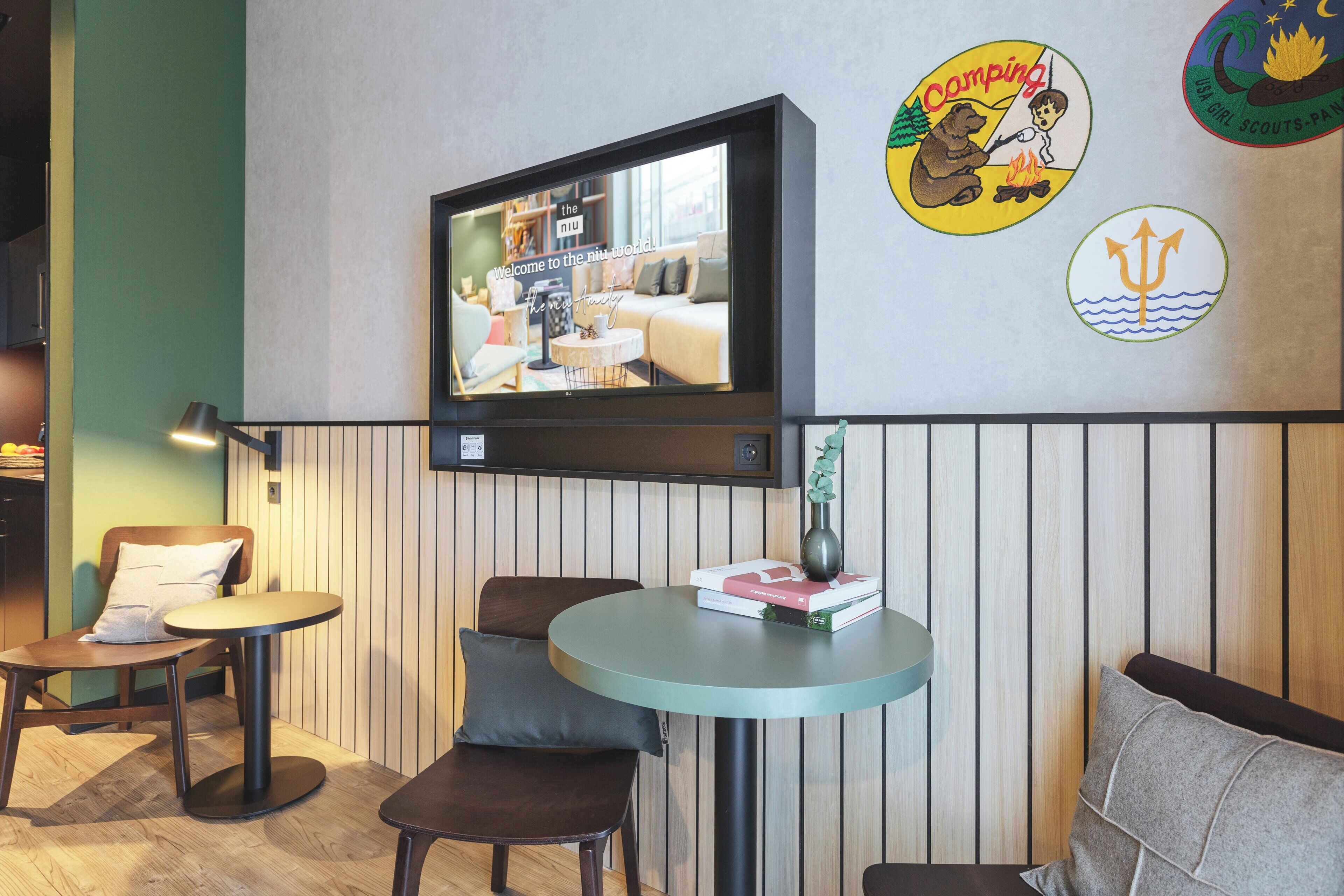 Ein modernes Zimmer mit an der Wand montiertem Fernseher, einem kleinen runden Tisch mit einem Buch, zwei Stühlen und einer Wand mit dekorativen Abzeichen.
