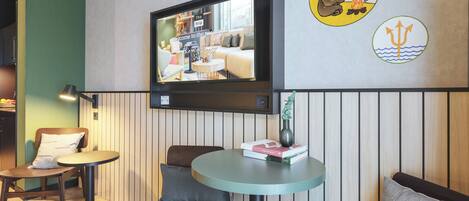 Une chambre moderne avec un téléviseur fixé au mur, une petite table ronde sur laquelle repose un livre, deux chaises et un mur décoratif orné d’écussons.