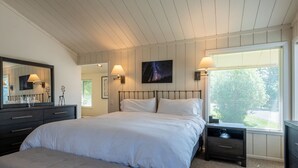 Un dormitorio con una cama grande, una mesita de noche, un aparador, un banco, un televisor montado en la pared y una ventana con persianas.