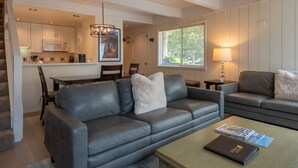 Una sala de estar moderna con un sofá de cuero gris, una mesa de centro con un libro, y un comedor con una mesa y sillas blancas.