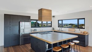 A modern kitchen with a large island, dark cabinetry, and a view of the outdoors through large windows. - Noble House - Absolutely Stunning House in Convenient Location! (Dalmeny)