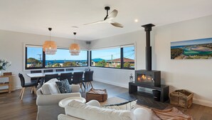 A modern living room with a sofa, dining table, and chairs, a fireplace, and large windows offering a view of the sea and hills. - Noble House - Absolutely Stunning House in Convenient Location! (Dalmeny)