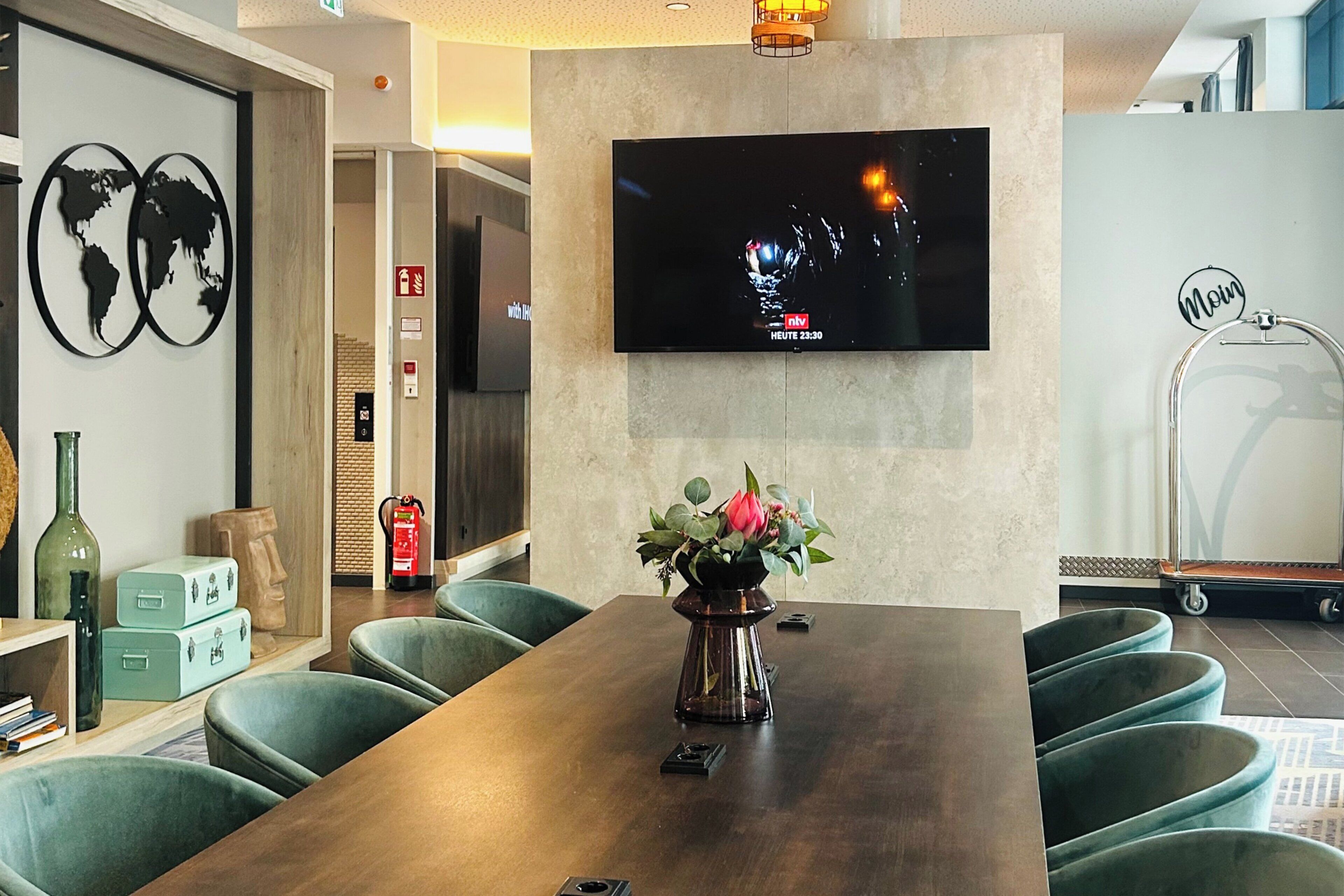 A modern hotel room with a large wooden dining table, green upholstered chairs, a flat-screen TV mounted on the wall, and a decorative vase with flowers on the table.