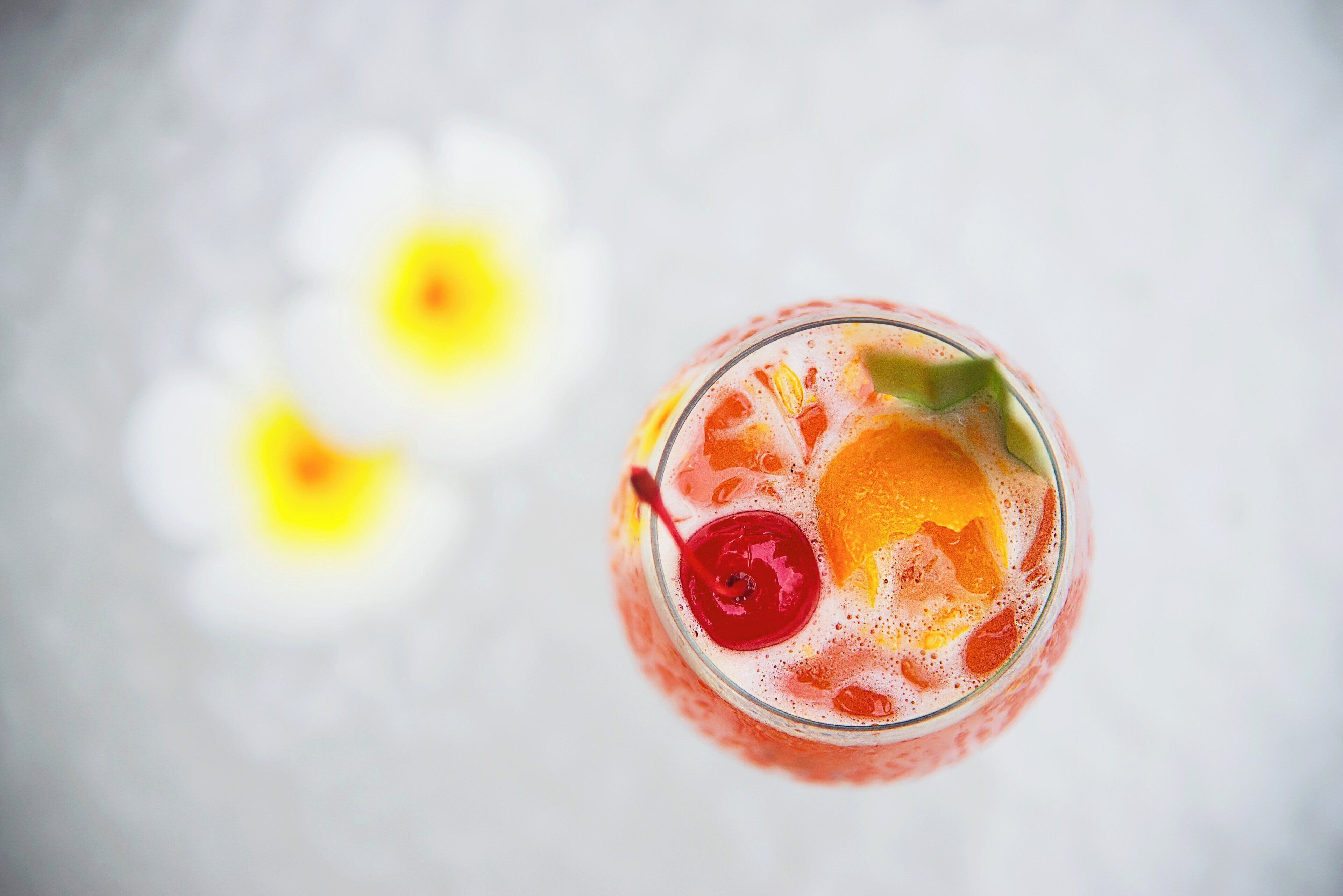 A glass of drink with fruit and a cherry on a white surface.