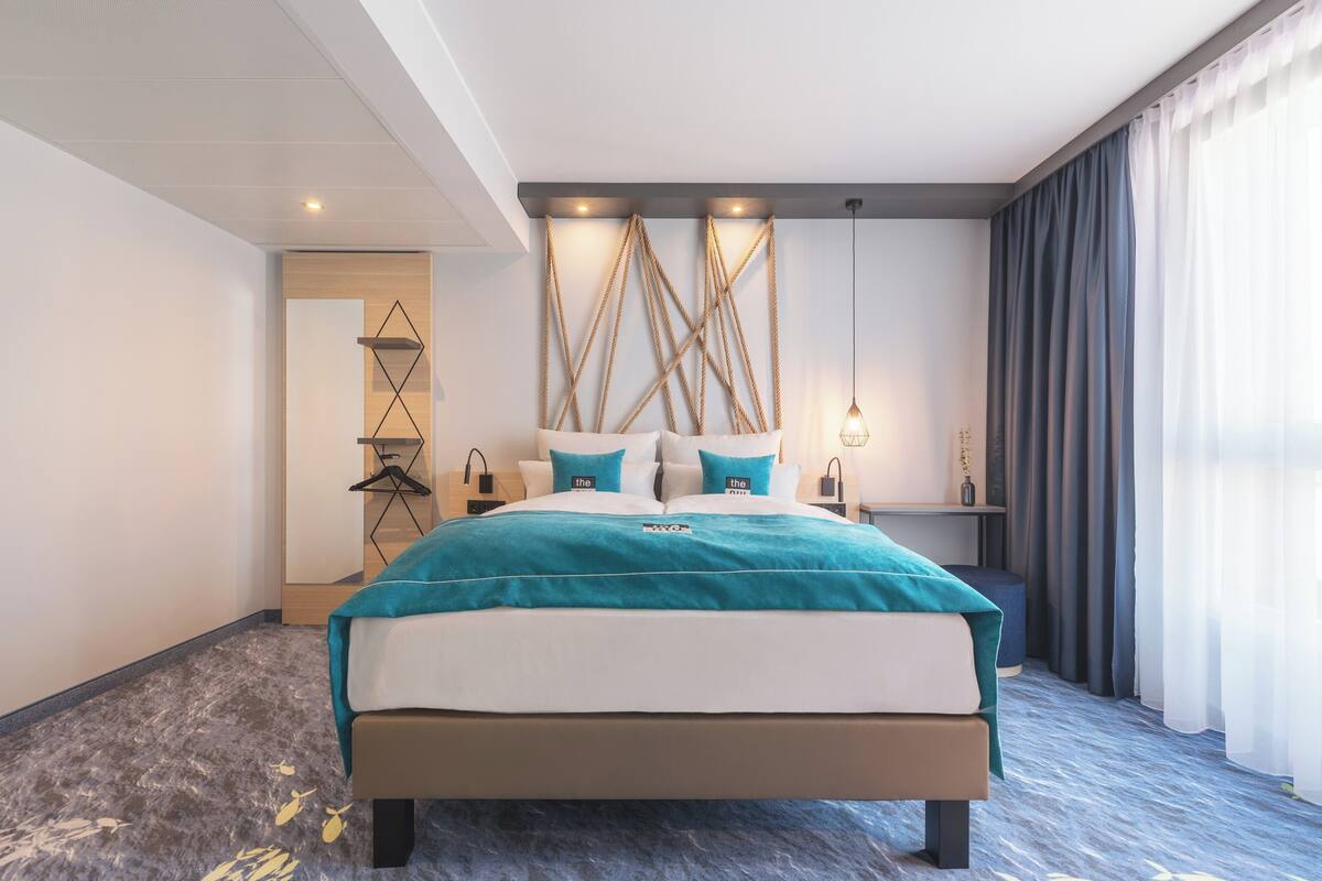Una habitación de hotel moderna con una cama grande, una mesita de noche, una lámpara colgante y vistas al exterior a través de cortinas translúcidas.