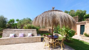Un patio exterior con suelo de piedra, una mesa redonda con sillas y un parasol de paja.