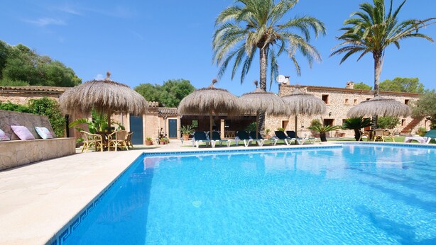 Piscina con sillas de descanso y sombrillas de paja, rodeada de palmeras y edificios de piedra.