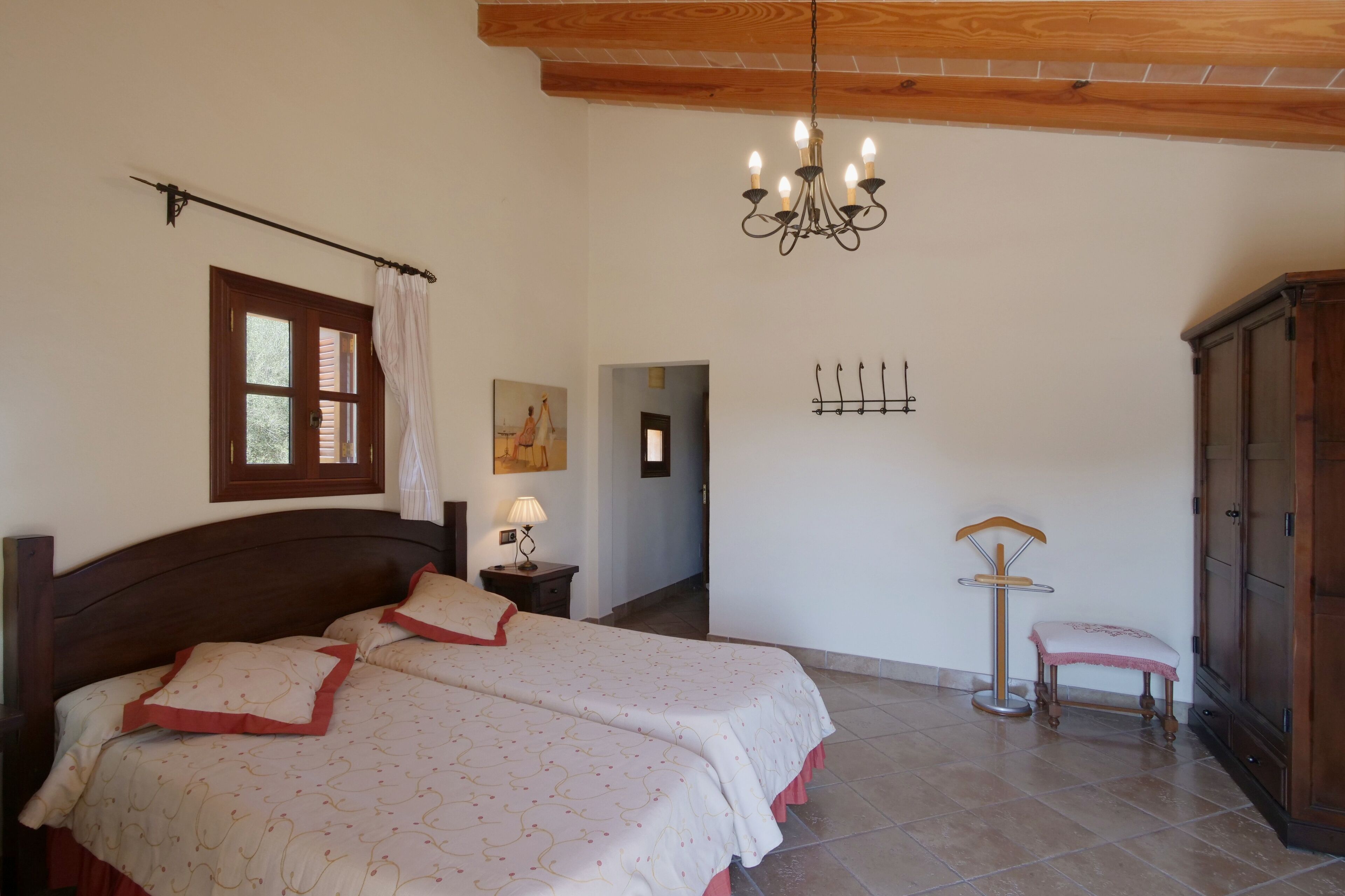 Un dormitorio con una cama grande, techo de madera, candelabro, ventana con cortinas, mesita de noche con lámpara, un cuadro en la pared y un armario.