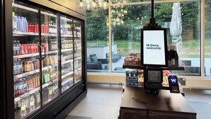 Un moderno vestíbulo de hotel con un amplio ventanal, una recepción con una pantalla digital que muestra “Hola, bienvenida/o”, y un expositor refrigerado de bebidas.