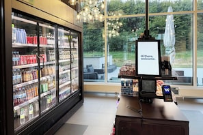 Un moderno vestíbulo de hotel con un amplio ventanal, una recepción con una pantalla digital que muestra “Hola, bienvenida/o”, y un expositor refrigerado de bebidas.