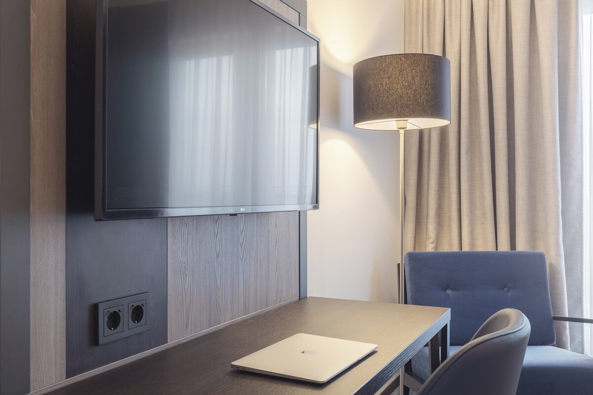 Habitación de hotel con televisión de pantalla plana en la pared, una lámpara, un ordenador portátil sobre una mesa y una silla.