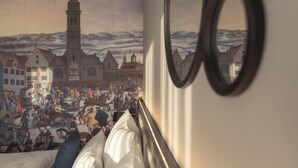 Una habitación con un gran mural que representa una escena histórica, una cama y un espejo que refleja parte del mural.