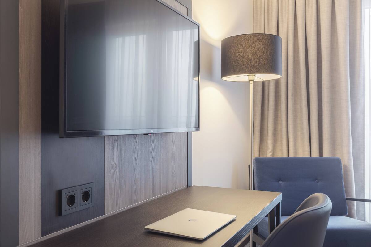 A hotel room with a flat-screen TV mounted on the wall, a lamp, a laptop on a table, and a chair.