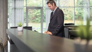 Un hombre con traje está de pie en la recepción de un moderno hotel, con grandes ventanales y una planta en maceta.