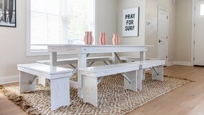 A white dining table with benches in a room with a large window, a rug, and a 'Pray for Surf' sign on the wall.