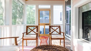 A covered porch with two wooden chairs, a small red table, and a woven ottoman.