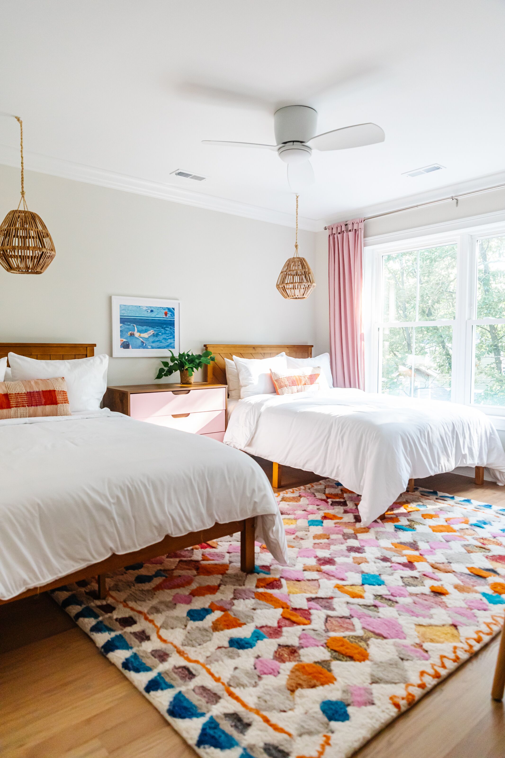 A bedroom with two beds, a ceiling fan, a window with pink curtains, and a colorful rug.
