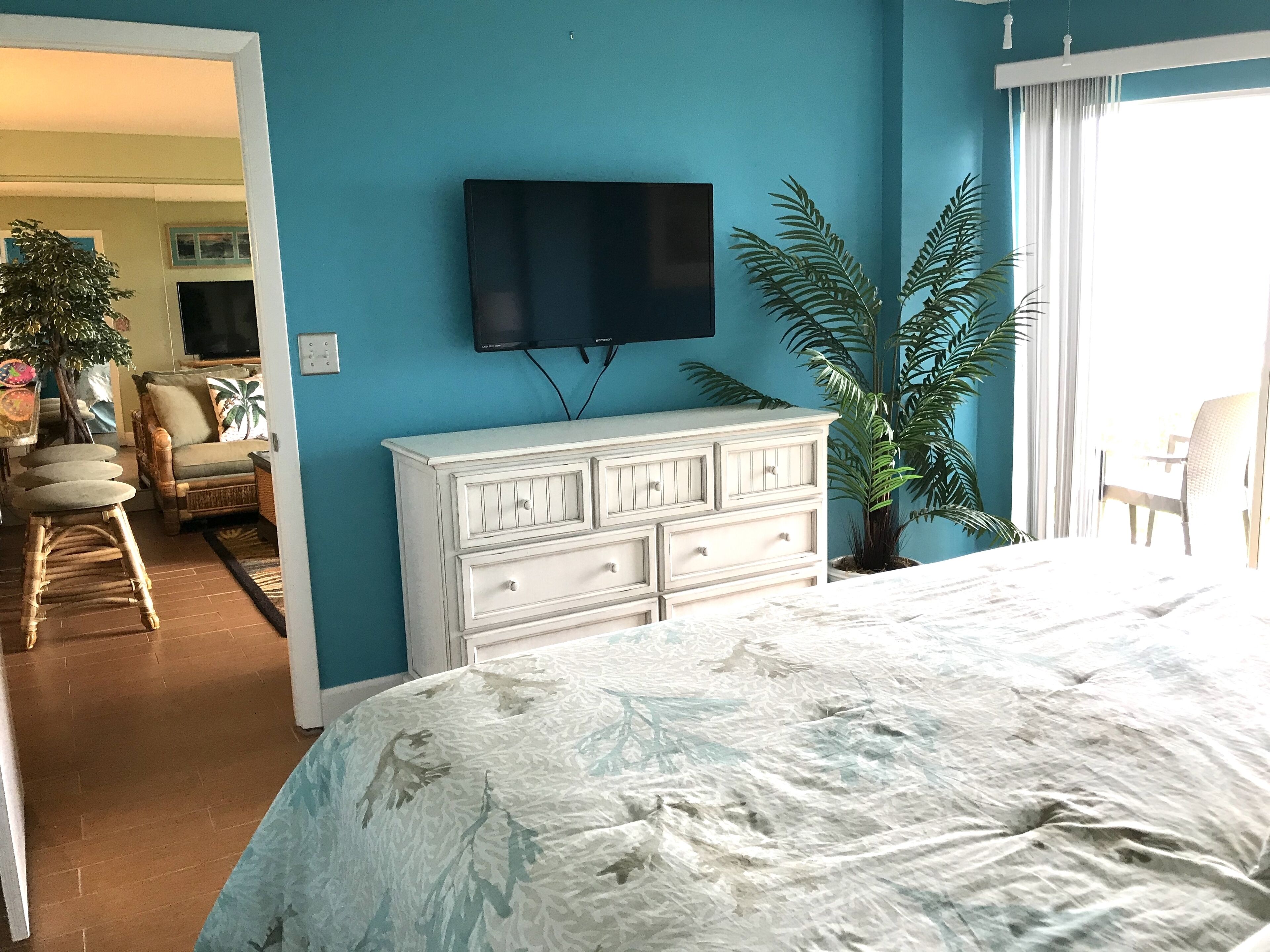 Un dormitorio con cama, cómoda, televisor en la pared y una planta grande.
