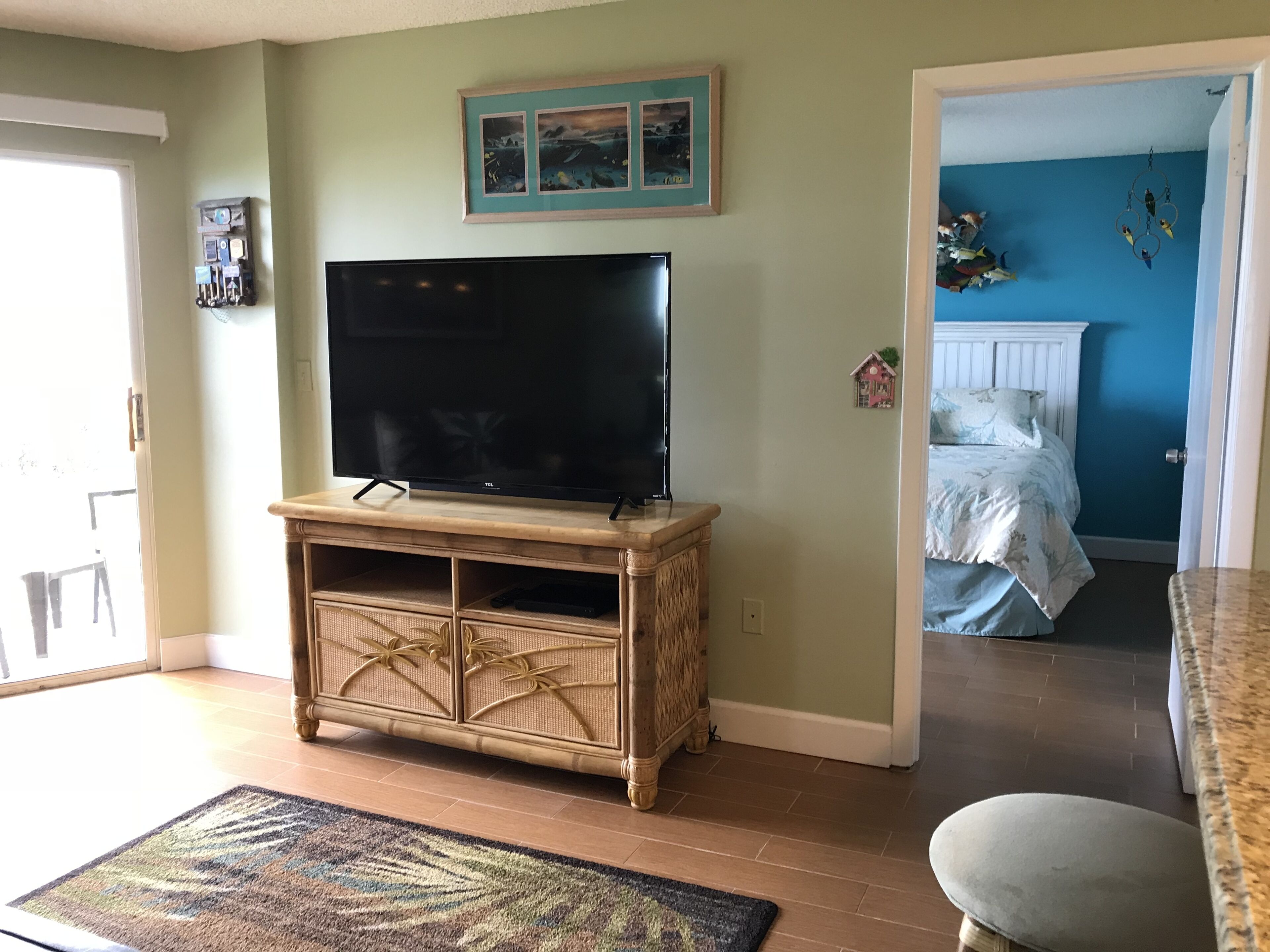 Un salon avec un téléviseur à écran plat posé sur un meuble en bois, un petit tapis et une porte donnant sur une chambre à coucher avec un lit et un mur bleu.