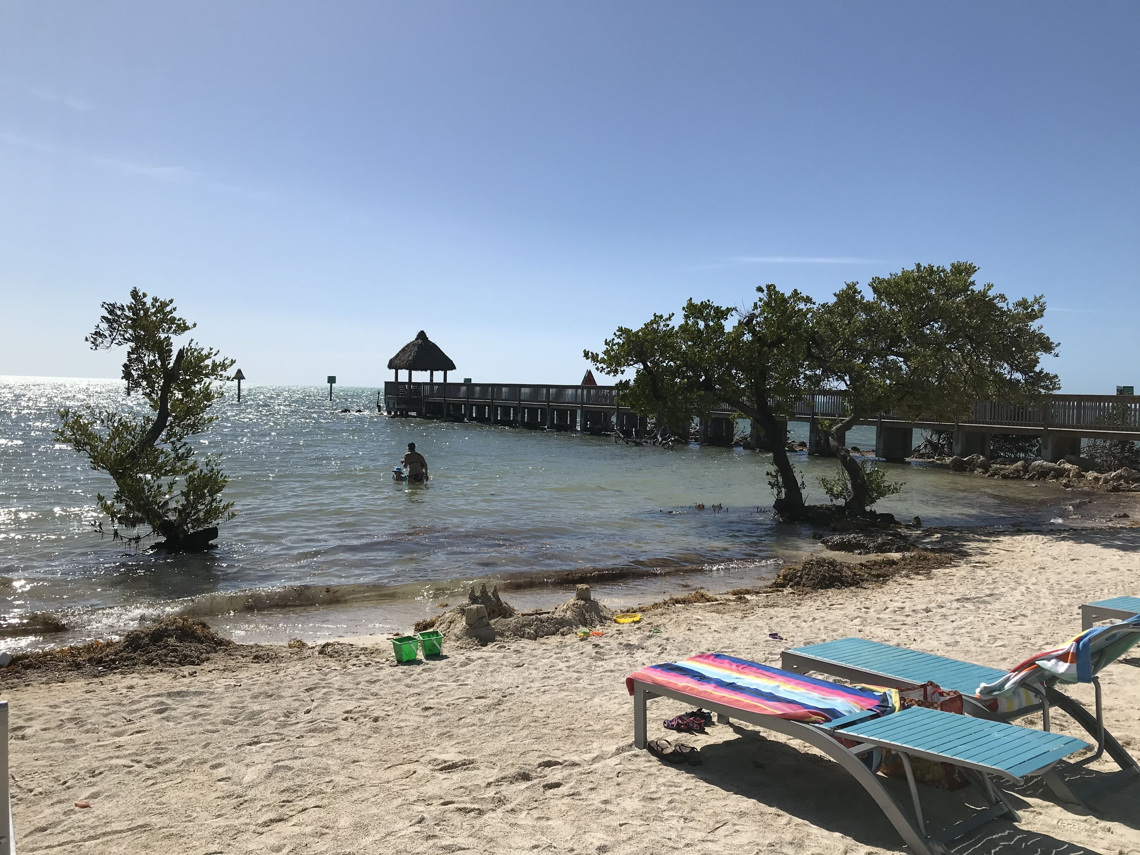 A beach with lounge chairs, a pier, and a thatched-roof structure.