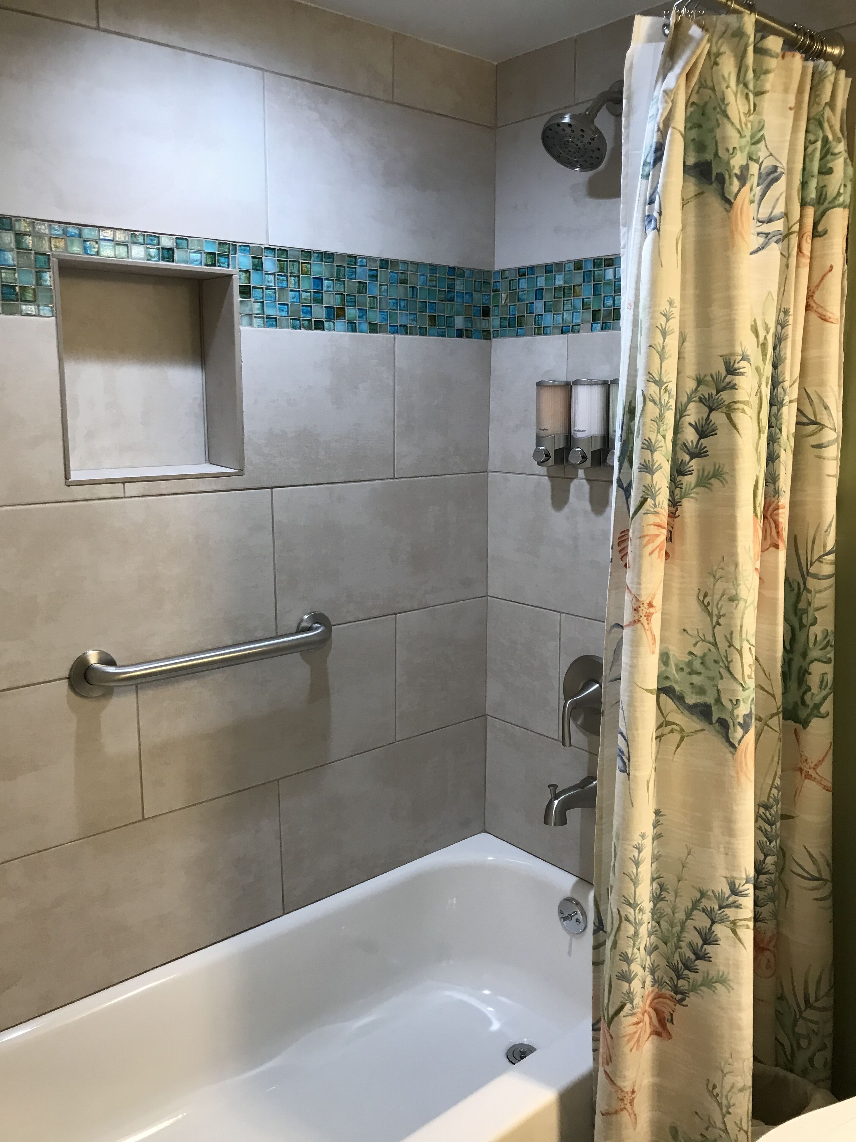 A bathroom with a bathtub, a shower with a rain showerhead, a handrail, and a small shelf.