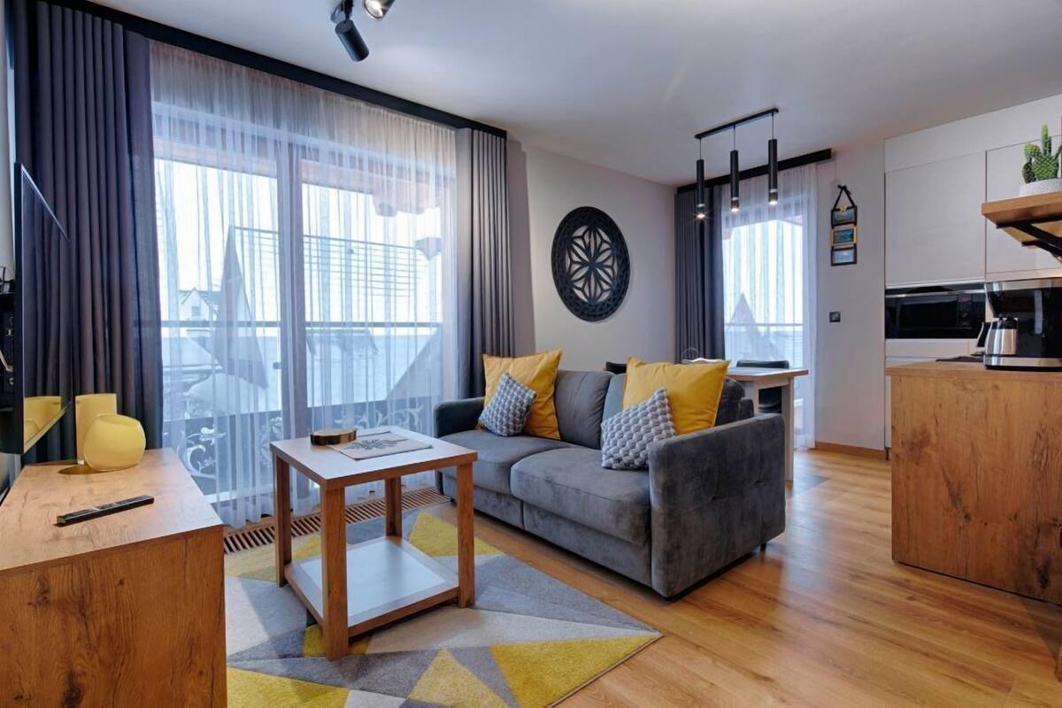 A modern living room with a grey sofa, wooden coffee table, and a large window with curtains.